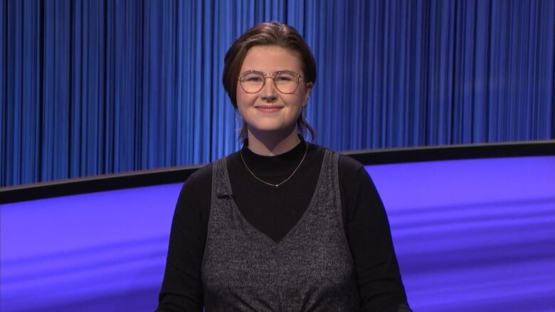 An image of Mattea Roach on the set of Jeopardy! They're wearing a long sleeve black shirt under a v-neck vest.