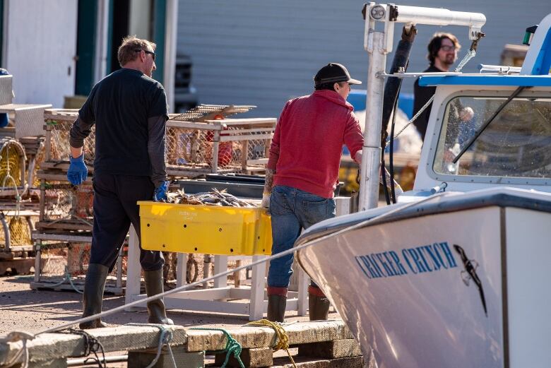 Two men carry large yellow tub of lobster on fishing wharf.