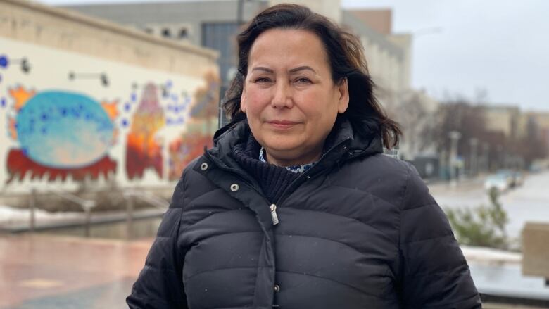 A woman with long black hair and wearing a black puffy winter jacket stands outside.