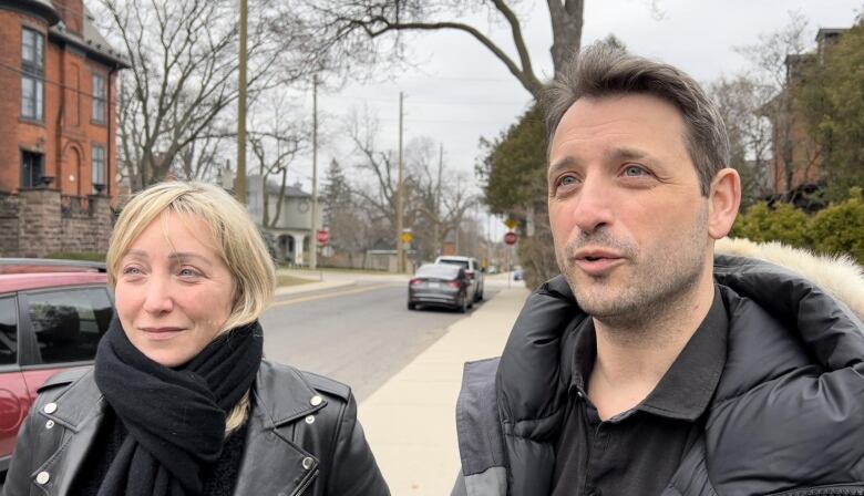 Two people in black coats stand beside each other on a street with tall trees and large houses.