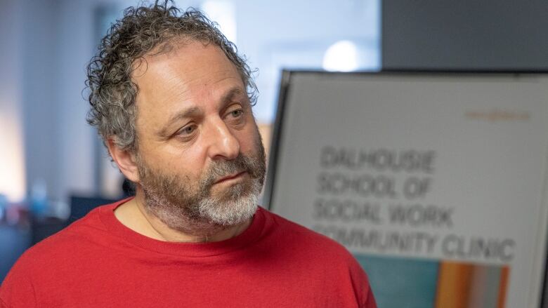 A man wearing a red shirt with salt-and-pepper curly hair stands in front of a sign that reads Dalhousie School of Social Work Community Clinic.