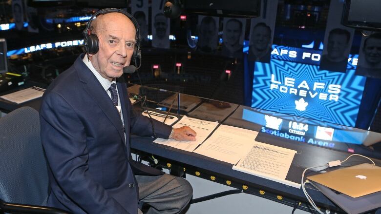 An older man wearing a suit sits in the broadcast booth at a hockey arena.