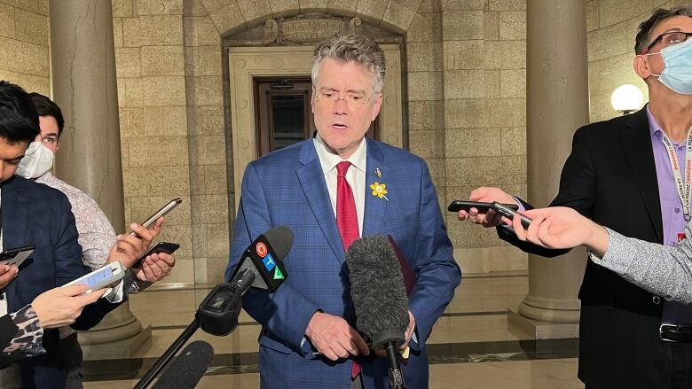 A politician stands inside a lobby of the Manitoba legislature while reporters standing on either side of him extend recording devices.