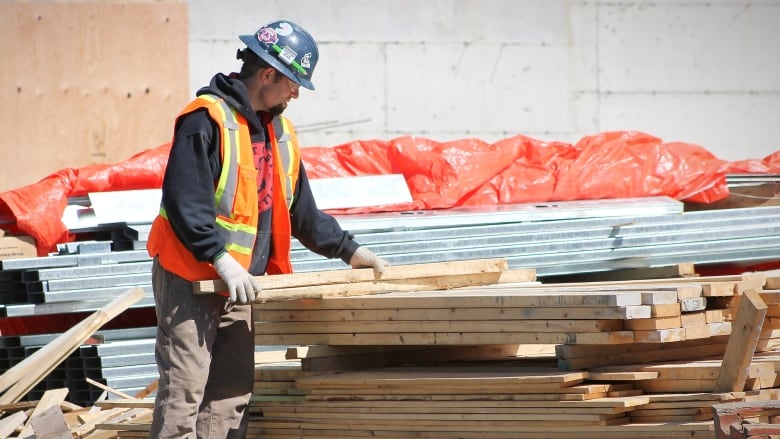 A man in construction gear and a hard hat handles a piece of wood that's on a pile of wood.