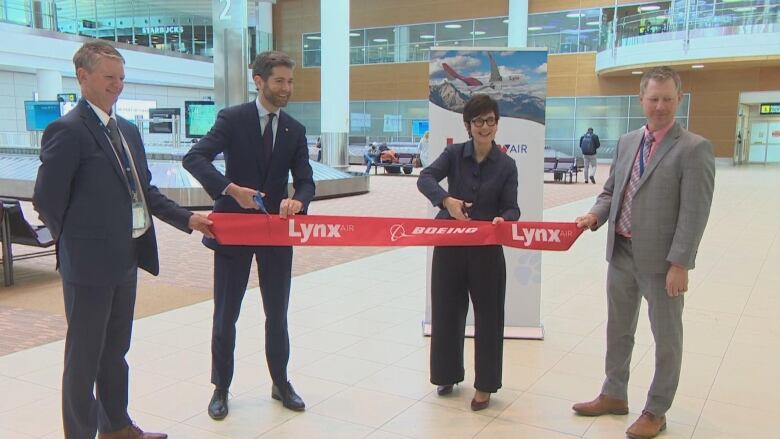 A woman in a dark pantsuit cuts a red ribbon in an airport.