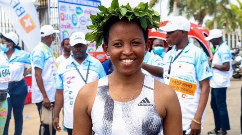 A woman in a white and black running uniform, and wearing a wreath of leaves on her head, smiles as she stands in front of a group of people in the background. 