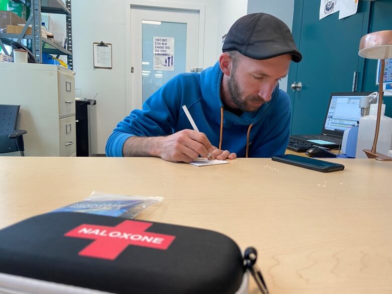 Jean-Franois Mary, executive director of CACTUS Montral, a harm reduction organization, sits at a table and writes. In the foreground, a naloxone kit can be seen on the table.