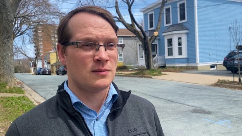 A white man with glasses looks to the side with a neutral expression. He is standing in a residential area on a sidewalk with homes behind him