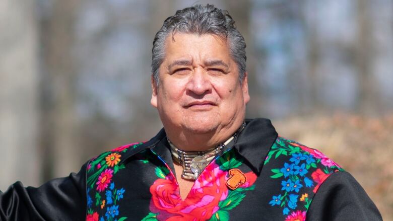 A man in a sweater with bright coloured flowers embroidered on it stands in a wooded area.