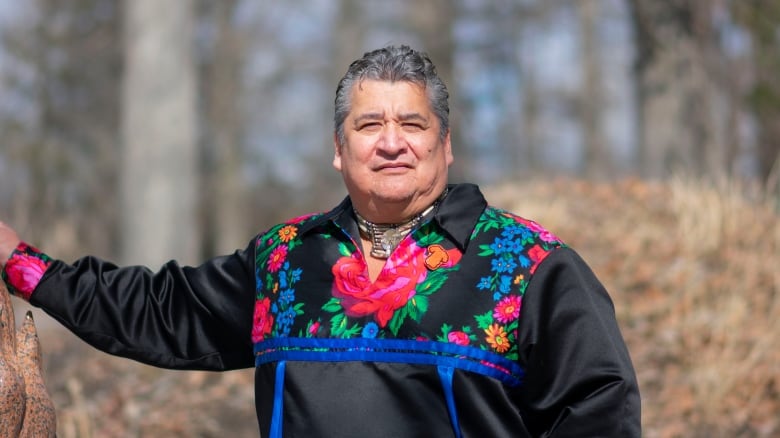 A man in a sweater with bright coloured flowers embroidered on it stands in a wooded area.