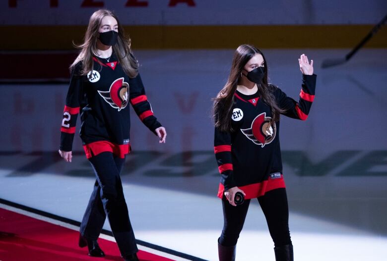 Two women in hockey sweaters walk on a carpet to centre ice.