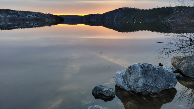 A sunset is reflected in calm river waters with rocks in the foreground.