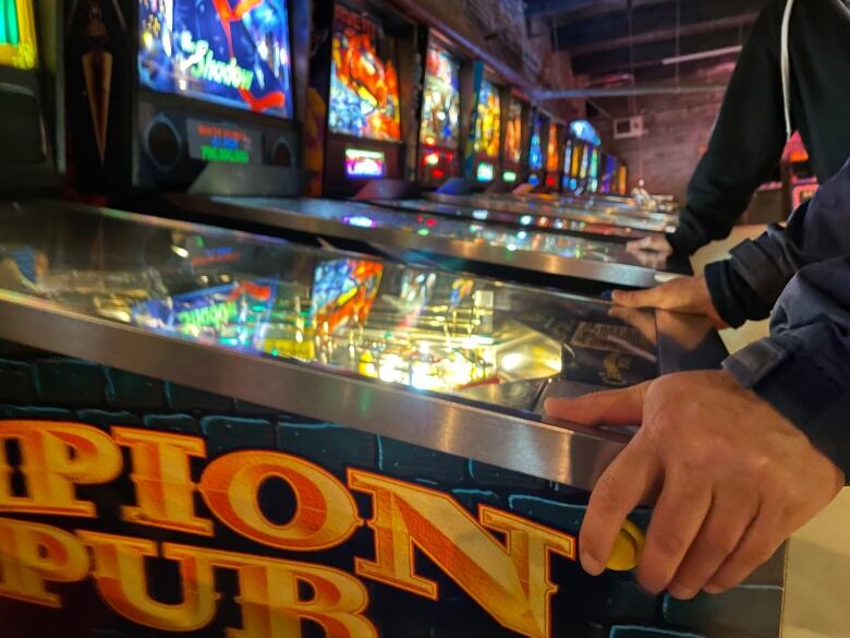 A person's two hands, resting on the edge of a pinball machine. There are other pinball machines in the background.