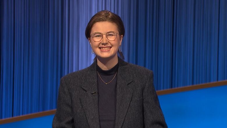 A person with hair styled in a pony tail wears a grey blazer with a grey shirt and glasses.