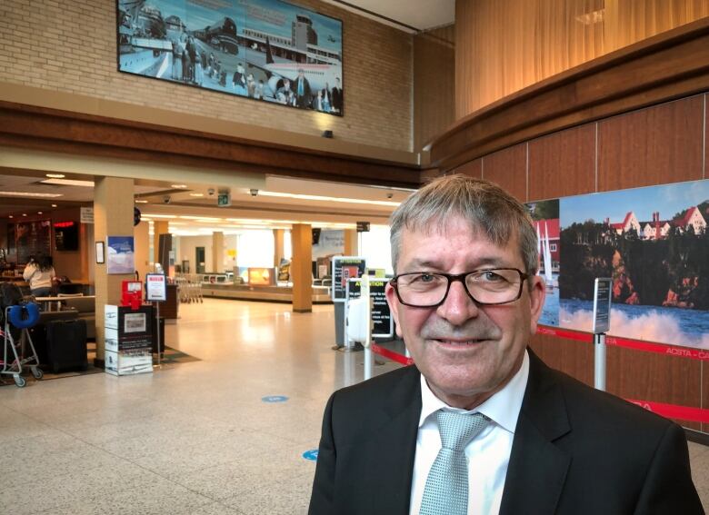 A man with glasses in a dark suit, white shirt and light blue tie stands in the airport waiting area.