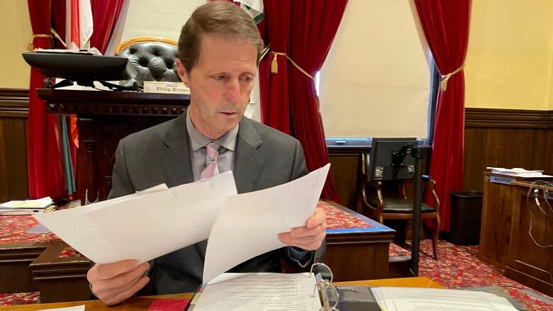 Man with short brown hair wearing grey suit jacket with tie sits at a desk in a city council room looking through papers.