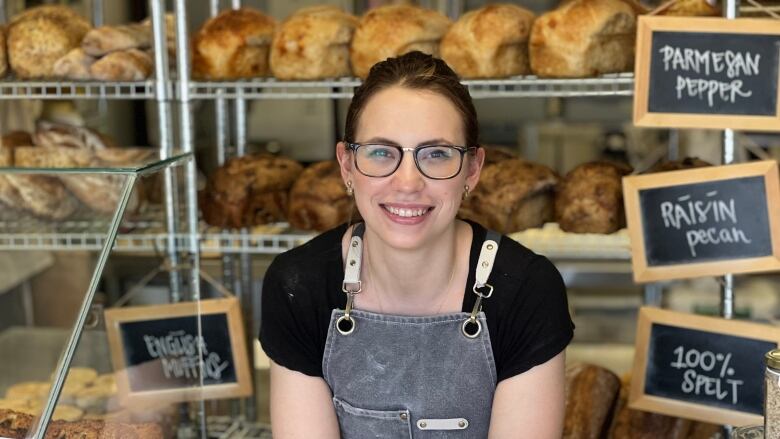 A woman in an apron stands in front of shelves filled with loaves of bread.