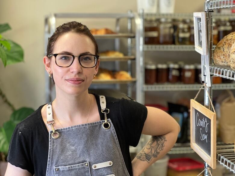 A woman in an apron stands in front of shelves of baking and jars of preserves.