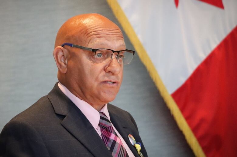 A man in a charcoal grey suit with glasses speaks to someone off camera with a Canadian flag in the background.