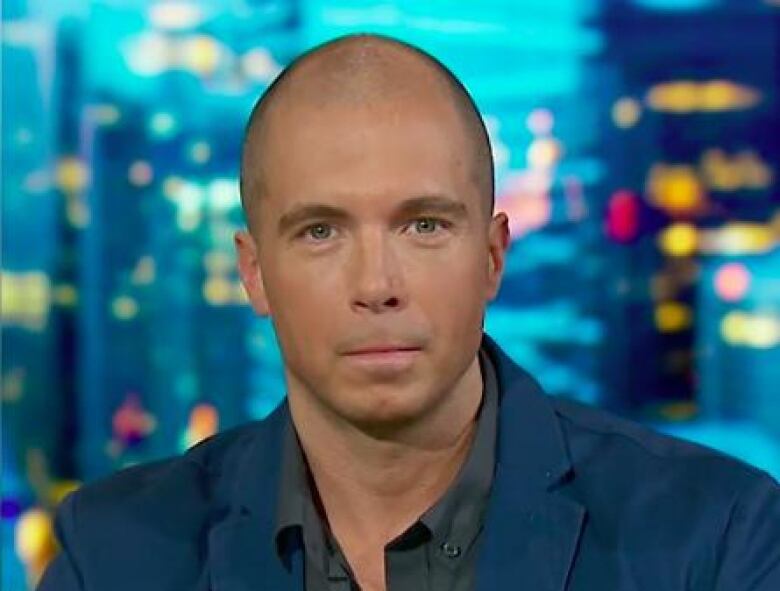 A balding man sitting in front of a window wears a navy blazer.