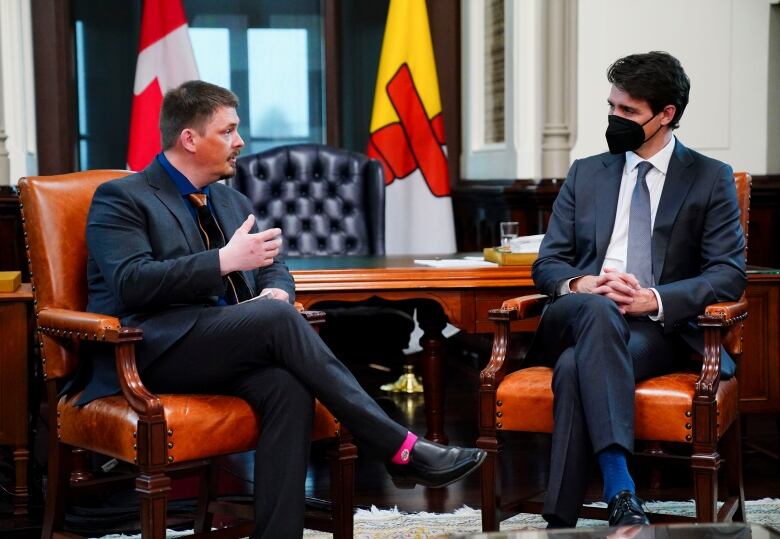 A man wearing no mask and a suit sits in a chair facing Prime Minister Justin Trudeau, who is wearing a mask and a navy blue suit.