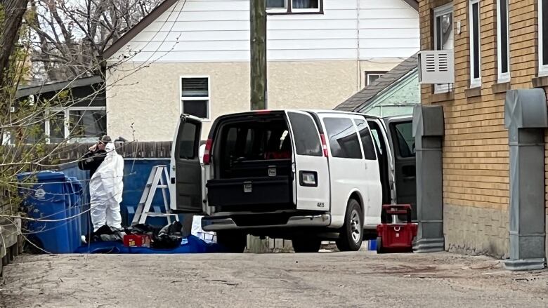 A police officer in a white zip-up suit stands near garbage and recycling bins next to a white van with the back open.