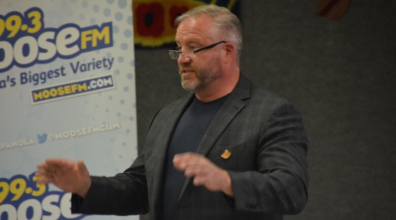 A man with grey hair and a beard wearing glasses and a blazer waves his hands while speaking to a crowd. 