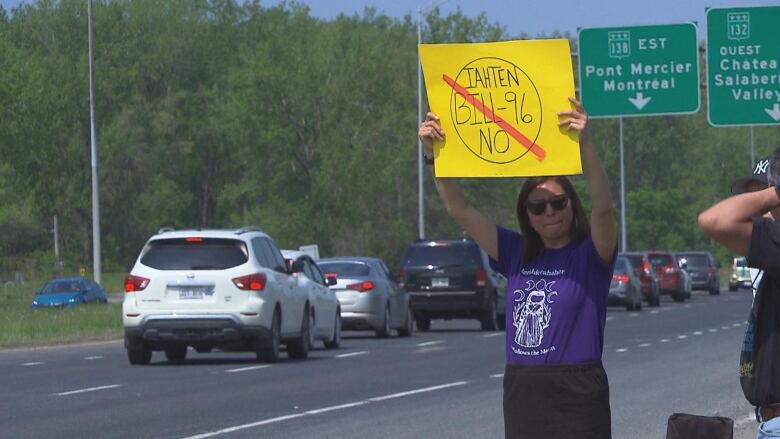 Woman standing by roadside holds yellow sign high that says Bill 96 no.