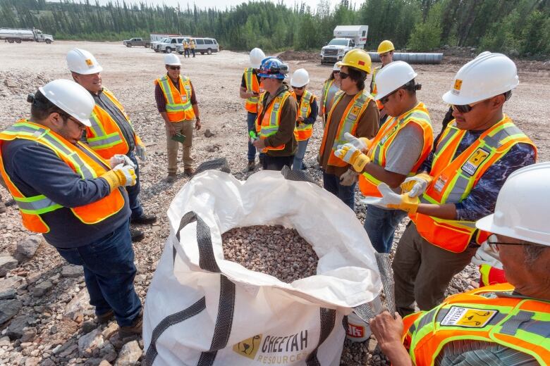 A group of people in safety vests and hardhats surround a big bag filled with rocks.