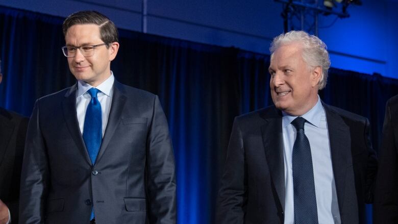 Two men in suits stand side by side on a stage. One is looking at the other, with an odd look on his face. 