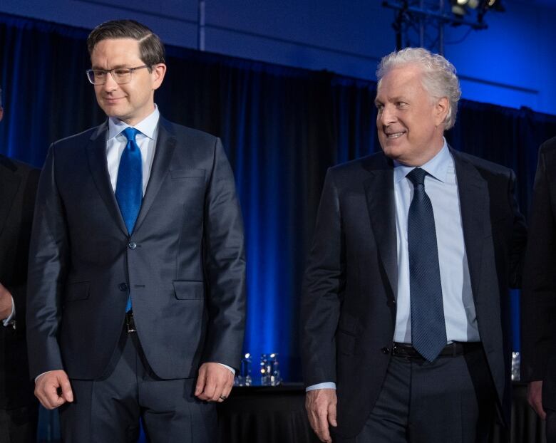 Two men in suits stand side by side on a stage. One is looking at the other, with an odd look on his face. 