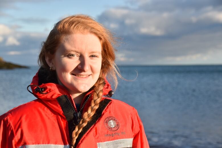 A woman in an orange jacket is standing outside with the ocean behind her.