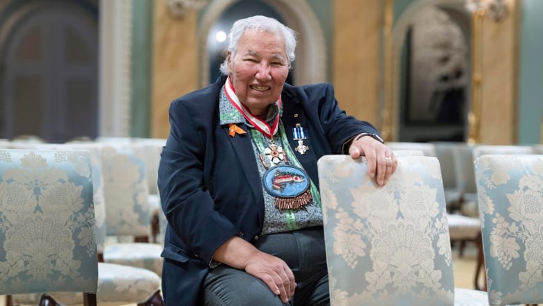 A man decorated in medals sits on a patterned chair while his left arm rests on the top of another chair in front of him. 