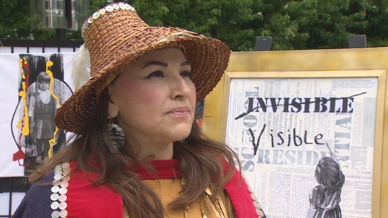 A woman in a conical Haida hat and regalia stands beside a poster that has the word  'Invisible' crossed out and replaced with the word 'visible.'