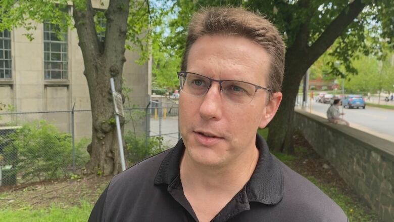 A man with brown hair and glasses wearing a chocolate coloured polo shirt stands in front of a stone wall with a tree near it.