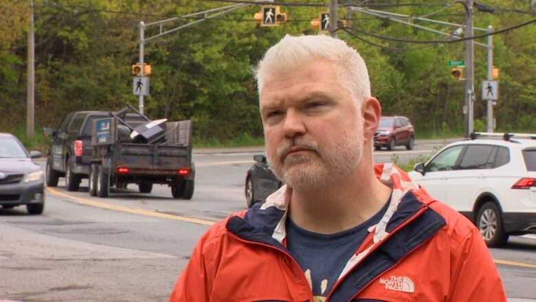 A white man with a white beard and orange jacket stands near an intersection