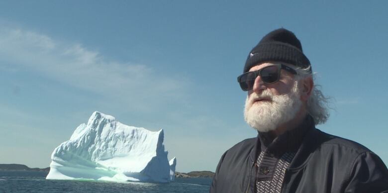 A man with a white bushy beard, wearing a black tuque and sunglasses, is interviewed next to a body of water, with a large glacier shown over his shoulder.