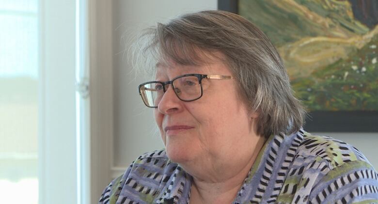 A woman wearing black-rimmed glasses and a patterned blouse sits for a conversation at a table. Behind her is a window and a framed photo.