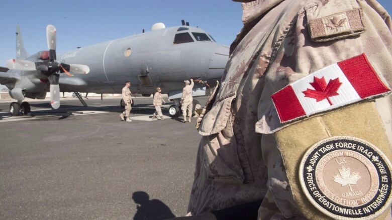 A soldier in desert camouflage stands in front of a plane on a tarmac.