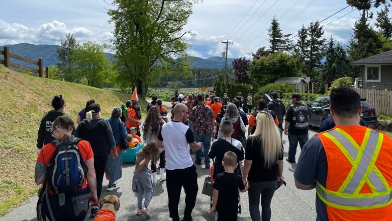 Marchers are seen on a side road. Some of them are wearing high visibility vests. All of them have their back to the camera. A child is visible holding a woman's hand.