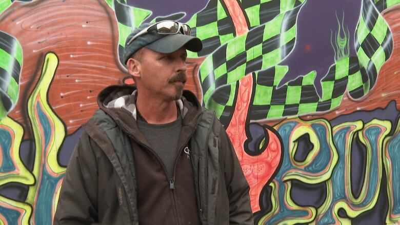 A man in a baseball cap stands in front of a wall covered in graffiti art.