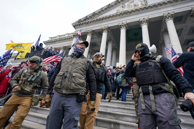 A large group of men  some masked and wearing tactical gear  stand on the steps of a government building. 