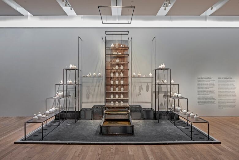 Photo of artwork installed in a gallery. A series of white vessels are arranged on stacked metal shelves.