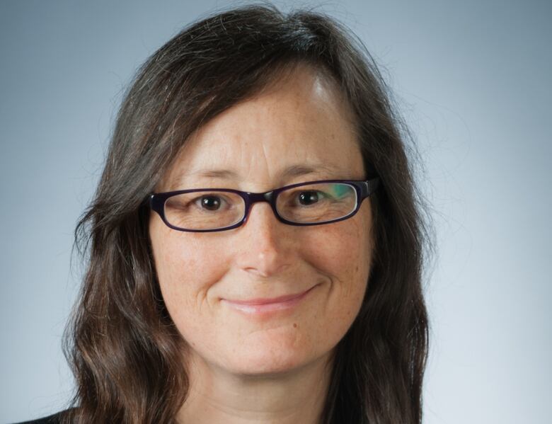 A woman with brown hair and glasses in front of a blue-grey background.