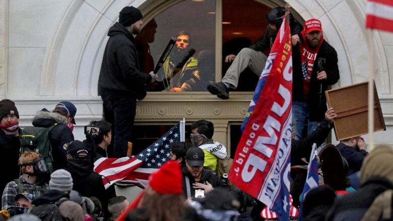 Crowds gathered in front of doorway, holding signs and U.S. flag, one man climbs through window, another holds a baseball bat.