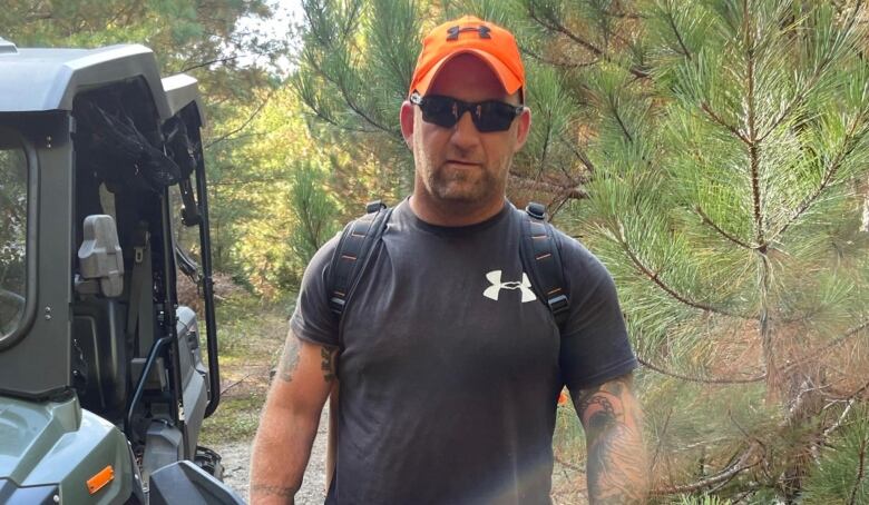 A man stands in a wooded area wearing an orange hat and sunglasses.