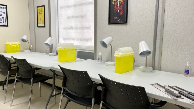 A row of chairs in front of a long counter that contains individual table lamps and safe disposal containers for used needles. 