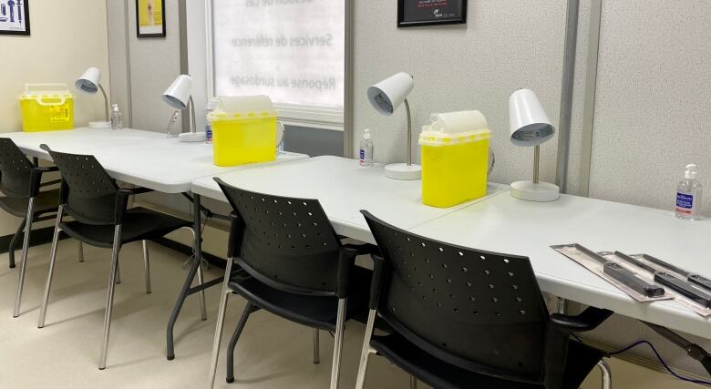 A row of chairs in front of a long counter that contains individual table lamps and safe disposal containers for used needles. 