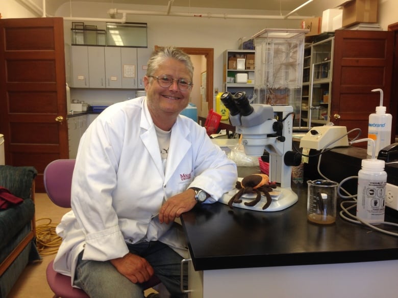 A smiling woman with glasses and short grey hair, wearing a lab coat, sits next to a microscope.