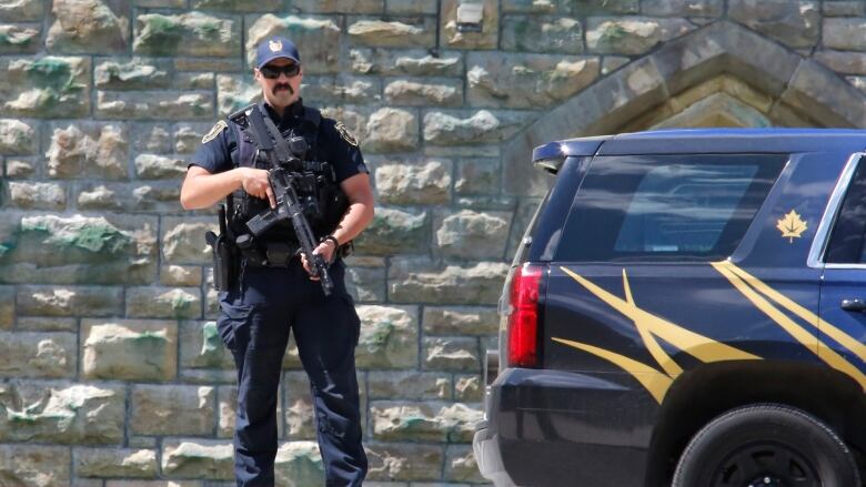 A law enforcement officer stands with a firearm beside a car.